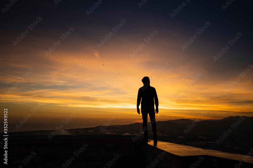 silueta de un hombre con capucha al atardecer con cielo de naranjas y violetas intensos silueta de un hombre con capucha al atardecer con cielo de naranjas y violetas intensos