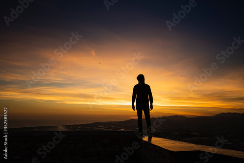 silueta de un hombre con capucha al atardecer con cielo de naranjas y violetas intensos © Ian