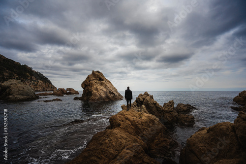 hombre de espalda parado sobre unas grandes rocas dentro del mar mediterráneo con el cielo nublado