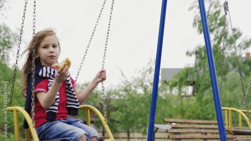 Little girl sitting on swing and eating hamburger on children playground. Summer windy day. The lockdown and quarantine ended. Little girl sitting on swing and eating hamburger on children playground. Summer windy day. The lockdown and quarantine ended.