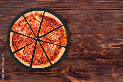 Sliced BBQ pizza with ham, bbq sauce, bacon and salami on a slate platter which is on wooden background, top view and space for text