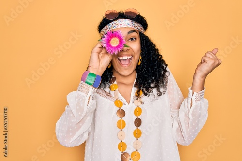 Young african american curly hippie woman wearing sunglasses holding pink flower on eye screaming proud and celebrating victory and success very excited, cheering emotion