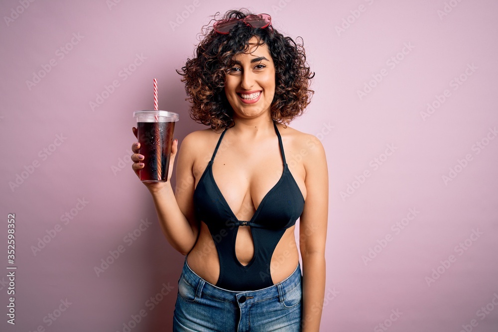 Beautiful arab woman on vacation wearing swimsuit drinking cola refreshment using straw with a happy face standing and smiling with a confident smile showing teeth Beautiful arab woman on vacation wearing swimsuit drinking cola refreshment using straw with a happy face standing and smiling with a confident smile showing teeth