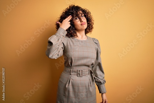 Beautiful arab business woman wearing dress and glasses standing over yellow background Shooting and killing oneself pointing hand and fingers to head like gun  suicide gesture.