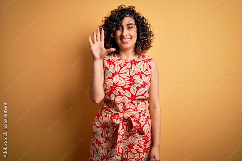 Young beautiful curly arab woman on vacation wearing summer floral dress and sunglasses showing and pointing up with fingers number five while smiling confident and happy. Young beautiful curly arab woman on vacation wearing summer floral dress and sunglasses showing and pointing up with fingers number five while smiling confident and happy.