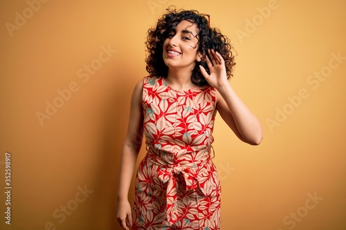 Young beautiful curly arab woman on vacation wearing summer floral dress and sunglasses smiling with hand over ear listening an hearing to rumor or gossip. Deafness concept.