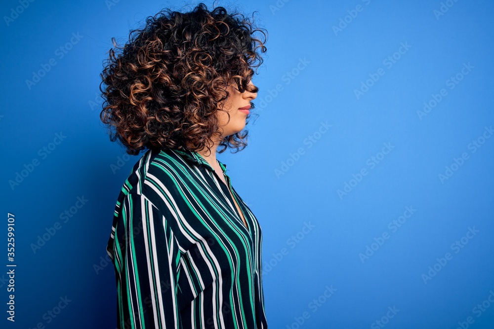 Young beautiful curly arab woman wearing striped shirt and glasses over blue background looking to side, relax profile pose with natural face with confident smile.