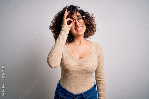 Young beautiful curly arab woman wearing casual t-shirt and glasses over white background doing ok gesture with hand smiling, eye looking through fingers with happy face.