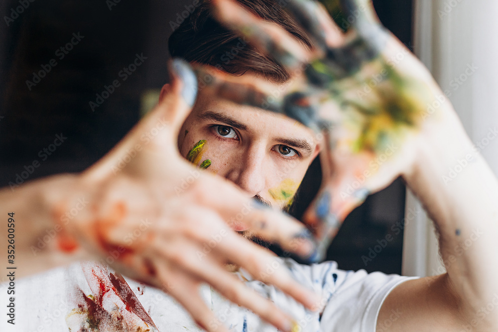 Portrait of a young creative artist, man wearing a white T-shirt with colored spots from paint posing for a photo in the process of creating a picture - obrazy, fototapety, plakaty 