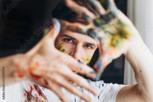 Portrait of a young creative artist, man wearing a white T-shirt with colored spots from paint posing for a photo in the process of creating a picture