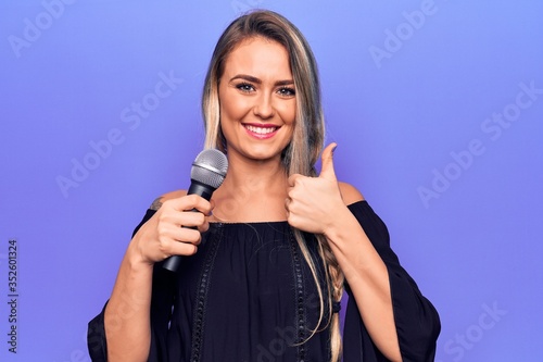 Young beautiful blonde singer woman singing using microphone over purple background smiling happy and positive, thumb up doing excellent and approval sign