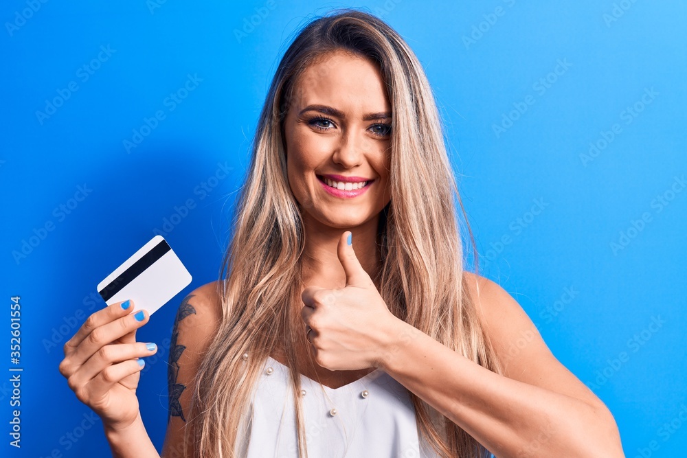 Young beautiful blonde woman holding credit card over isolated blue background smiling happy and positive, thumb up doing excellent and approval sign Young beautiful blonde woman holding credit card over isolated blue background smiling happy and positive, thumb up doing excellent and approval sign