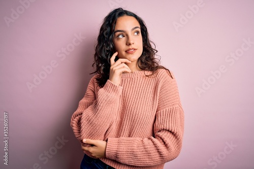 Young beautiful woman with curly hair wearing casual sweater over isolated pink background with hand on chin thinking about question, pensive expression. Smiling and thoughtful face. Doubt concept. © Krakenimages.com
