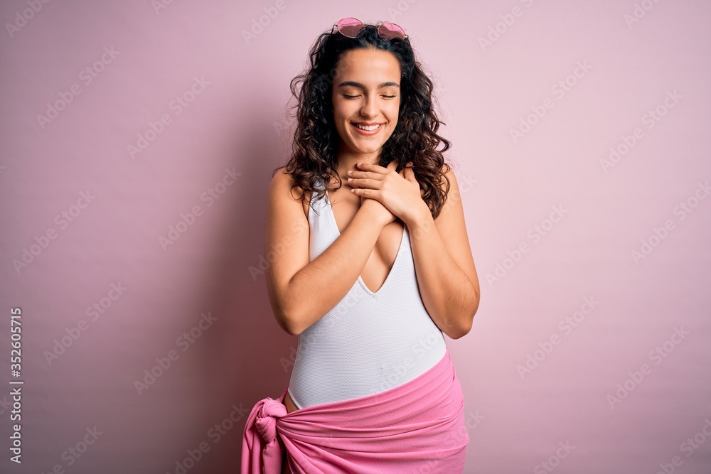 Beautiful woman with curly hair on vacation wearing white swimsuit over pink background smiling with hands on chest with closed eyes and grateful gesture on face. Health concept. Beautiful woman with curly hair on vacation wearing white swimsuit over pink background smiling with hands on chest with closed eyes and grateful gesture on face. Health concept.