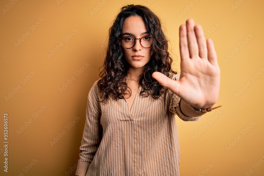 Beautiful woman with curly hair wearing striped shirt and glasses over yellow background doing stop sing with palm of the hand. Warning expression with negative and serious gesture on the face. Beautiful woman with curly hair wearing striped shirt and glasses over yellow background doing stop sing with palm of the hand. Warning expression with negative and serious gesture on the face.