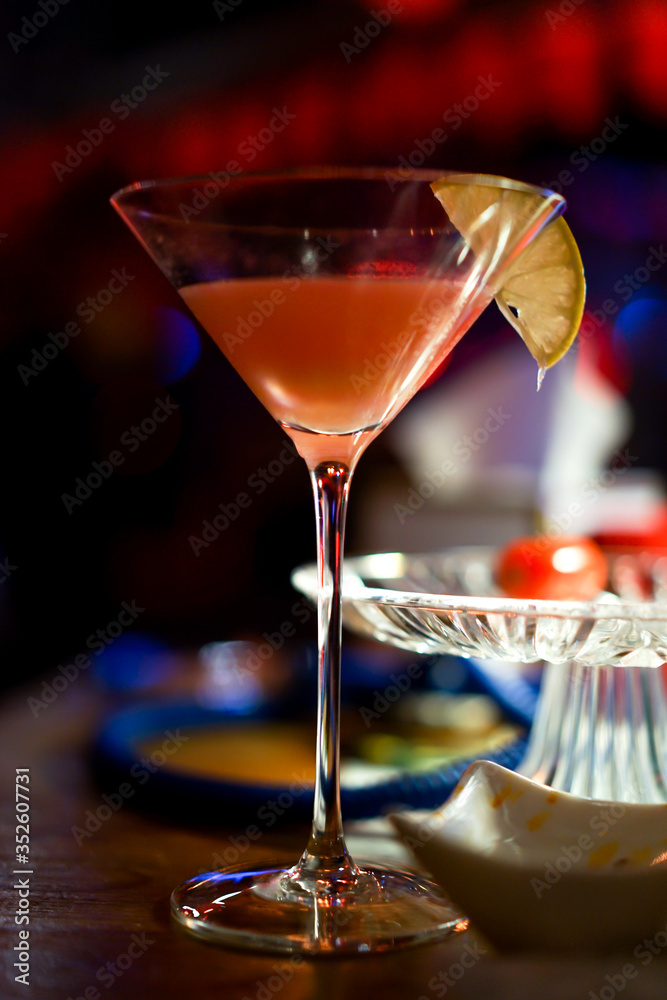 Tall martini glass with a pink cocktail and a slice of lemon on the table in the dark night club, selective focus