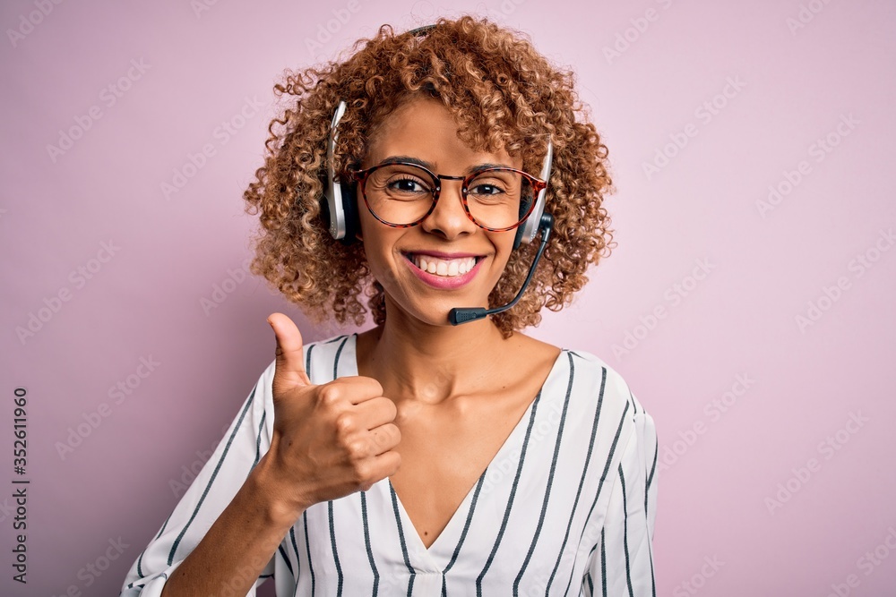 African american curly call center agent woman working using headset over pink background doing happy thumbs up gesture with hand. Approving expression looking at the camera showing success. African american curly call center agent woman working using headset over pink background doing happy thumbs up gesture with hand. Approving expression looking at the camera showing success.