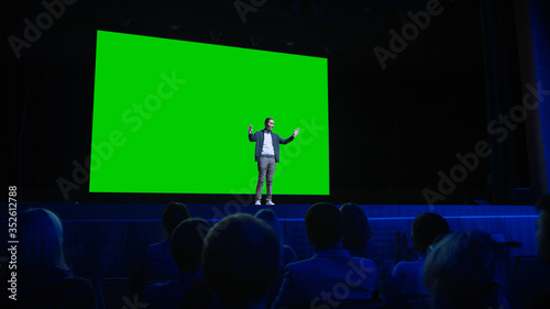 On Stage, Keynote Speaker Presents New Product to the Audience, Behind Him Movie Theater with Green Screen, Mock-up, Chroma Key. Business Live Event or Device Reveal photo