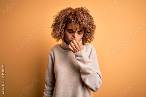 Young beautiful african american woman wearing turtleneck sweater over yellow background smelling something stinky and disgusting, intolerable smell, holding breath with fingers on nose. Bad smell © Krakenimages.com