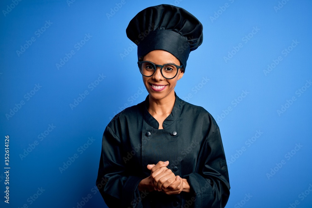 Young african american chef woman wearing cooker uniform and hat over blue background with hands together and crossed fingers smiling relaxed and cheerful. Success and optimistic Young african american chef woman wearing cooker uniform and hat over blue background with hands together and crossed fingers smiling relaxed and cheerful. Success and optimistic