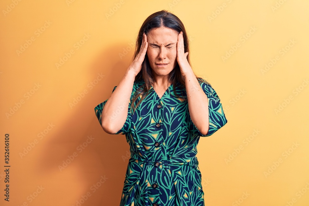 Young beautiful brunette woman wearing casual floral dress standing over yellow background with hand on head for pain in head because stress. Suffering migraine. Young beautiful brunette woman wearing casual floral dress standing over yellow background with hand on head for pain in head because stress. Suffering migraine.