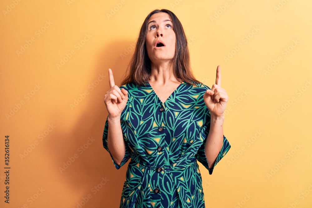 Young beautiful brunette woman wearing casual floral dress standing over yellow background amazed and surprised looking up and pointing with fingers and raised arms. Young beautiful brunette woman wearing casual floral dress standing over yellow background amazed and surprised looking up and pointing with fingers and raised arms.