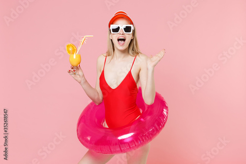 Surprised woman in red one-piece swimsuit cap glasses with swim inflatable ring isolated on pink background. People summer vacation rest lifestyle concept. Hold glass with cocktail spreading hands.