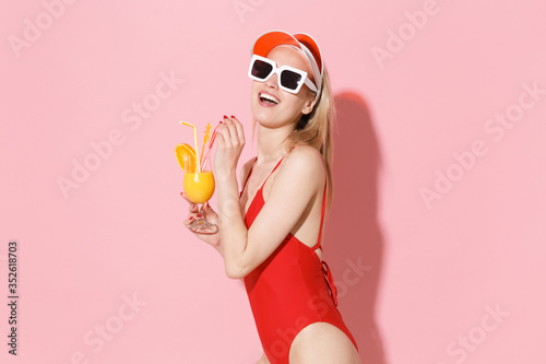 Side view of cheerful young blonde woman girl in red one-piece swimsuit cap sunglasses isolated on pink wall background studio. People summer vacation rest lifestyle concept. Hold glass with cocktail.