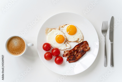 Homemade breakfast with fried eggs, bacon, fresh cherry tomatoes and a cup of coffee on the white table. Top view.