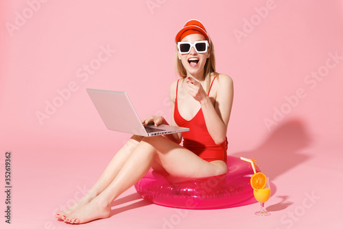 Excited young woman in red one-piece swimsuit cap sunglasses isolated on pink background. People summer vacation rest concept. Sit in swim inflatable ring, work on laptop pc computer, point on camera.