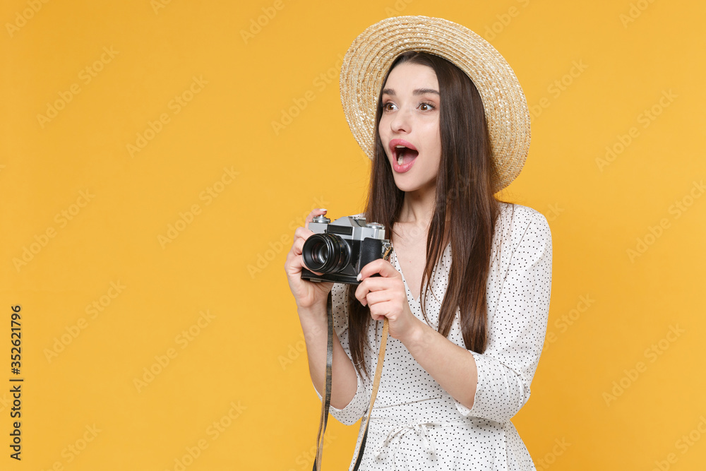 Shocked young brunette woman girl in white dress hat posing isolated on yellow wall background in studio. People lifestyle concept. Mock up copy space. Taking pictures with retro vintage photo camera.