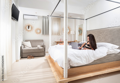 Young woman with laptop laying on white bed in modern design bedroom interior with light gray and natural wood furniture © GeorgeTsamakdas