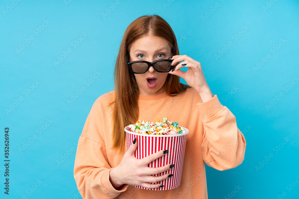 Young redhead woman over isolated background surprised with 3d glasses and holding a big bucket of popcorns