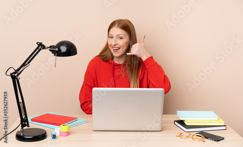 Young student woman in a workplace with a laptop making phone gesture. Call me back sign photo