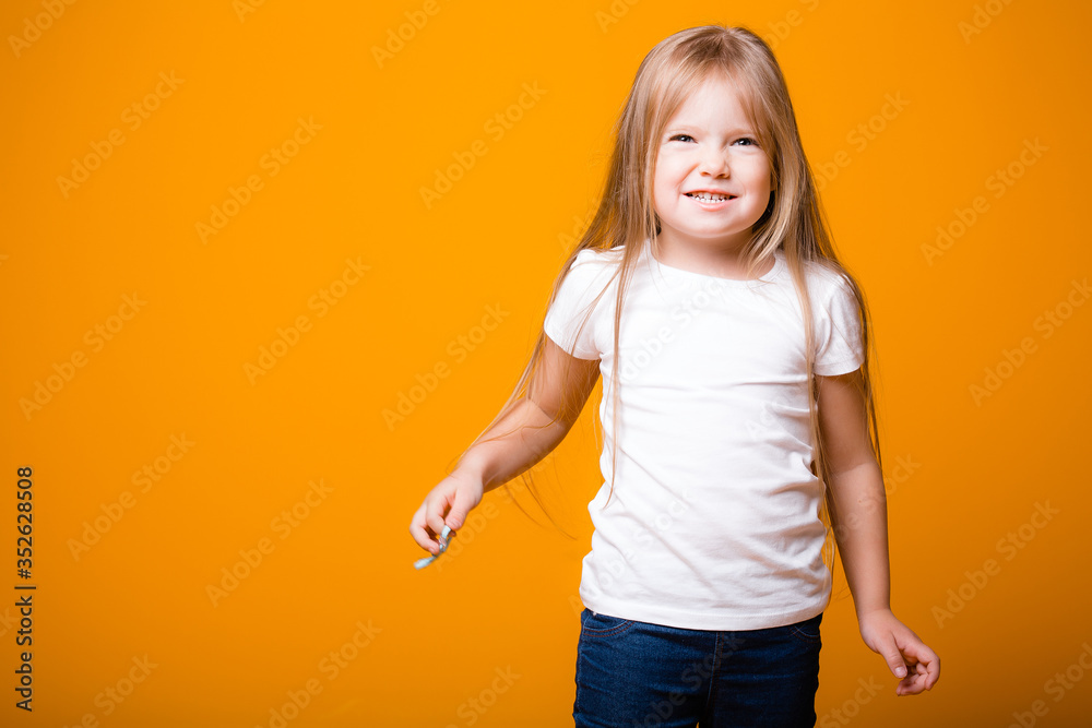 A child girl with long blond hair is holding a hair accessory in her hands. standing on an orange background A child girl with long blond hair is holding a hair accessory in her hands. standing on an orange background