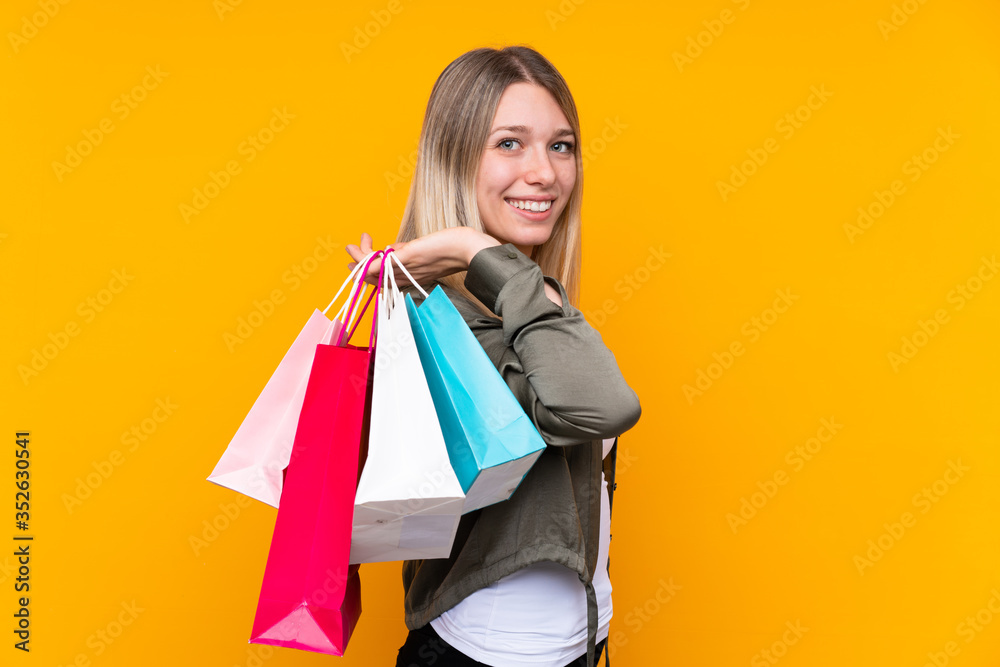 Young blonde woman over isolated yellow background holding shopping bags and smiling