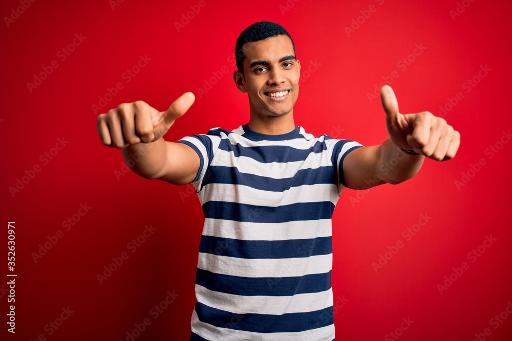 Handsome african american man wearing casual striped t-shirt standing over red background approving doing positive gesture with hand, thumbs up smiling and happy for success. Winner gesture. Handsome african american man wearing casual striped t-shirt standing over red background approving doing positive gesture with hand, thumbs up smiling and happy for success. Winner gesture.