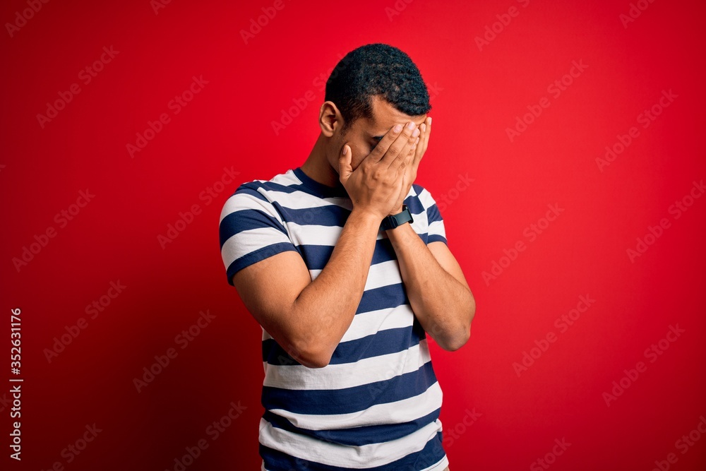 Handsome african american man wearing casual striped t-shirt standing over red background with sad expression covering face with hands while crying. Depression concept. Handsome african american man wearing casual striped t-shirt standing over red background with sad expression covering face with hands while crying. Depression concept.