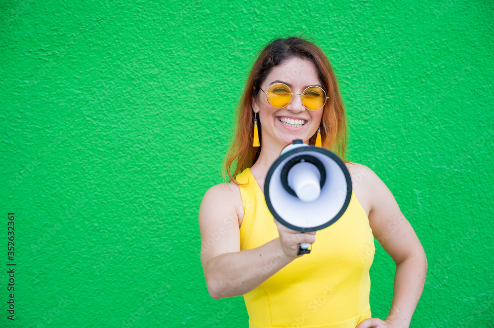 Fototapeta premium Happy woman in a yellow dress glasses and earrings on a green background shouting into a megaphone. Portrait of a girl holding a loudspeaker. Lady with a perfect snow-white smile.