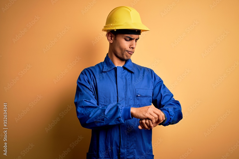 Young handsome african american worker man wearing blue uniform and security helmet Checking the time on wrist watch, relaxed and confident