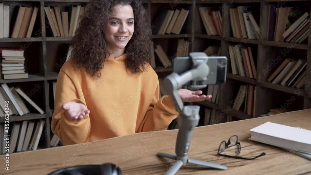 Smiling hispanic gen z teen girl social media channel blogger content creator recording vlog video tutorial on smartphone cam in library. School student vlogger talk look at mobile phone shoot blog.