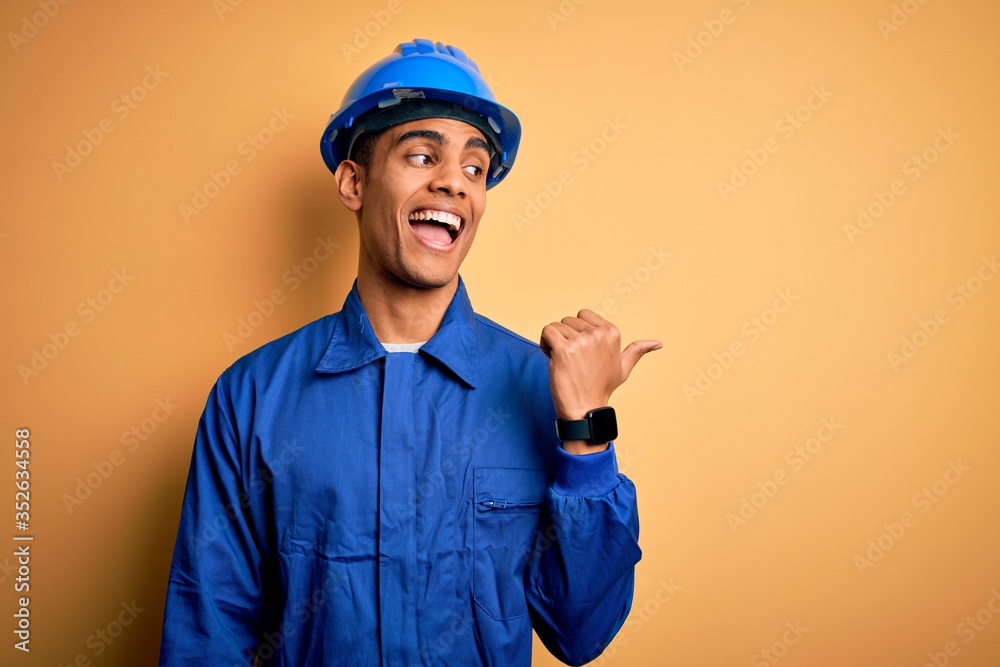 Young handsome african american worker man wearing blue uniform and security helmet smiling with happy face looking and pointing to the side with thumb up.
