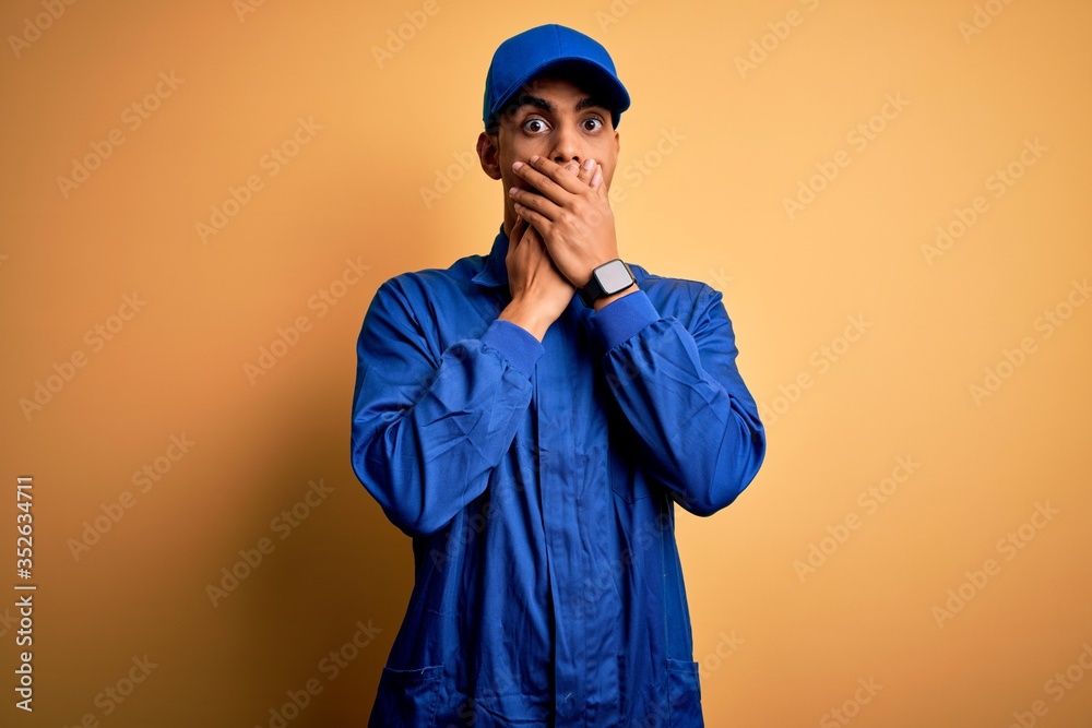 Young african american mechanic man wearing blue uniform and cap over yellow background shocked covering mouth with hands for mistake. Secret concept.