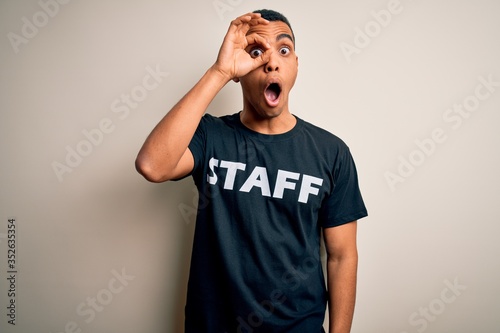 Young handsome african american worker man wearing staff uniform over white background doing ok gesture shocked with surprised face, eye looking through fingers. Unbelieving expression.