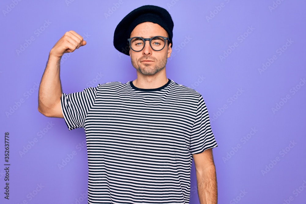 Handsome man with blue eyes wearing striped t-shirt and french beret over purple background Strong person showing arm muscle, confident and proud of power Handsome man with blue eyes wearing striped t-shirt and french beret over purple background Strong person showing arm muscle, confident and proud of power