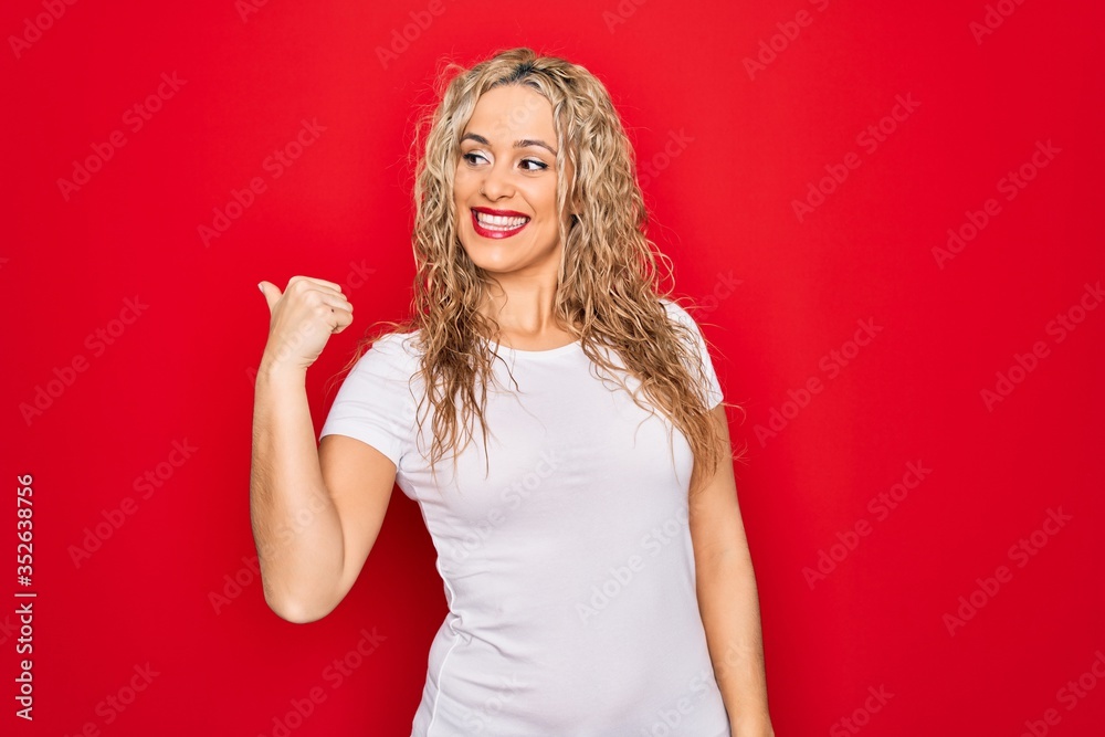 Young beautiful blonde woman wearing casual t-shirt standing over isolated red background smiling with happy face looking and pointing to the side with thumb up. Young beautiful blonde woman wearing casual t-shirt standing over isolated red background smiling with happy face looking and pointing to the side with thumb up.
