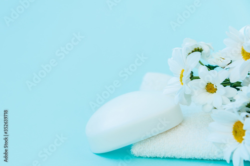 On a white Terry towel is a piece of handmade soap. Beautiful flowers of white daisies lie nearby. The background of the image is blue. There is space for text.