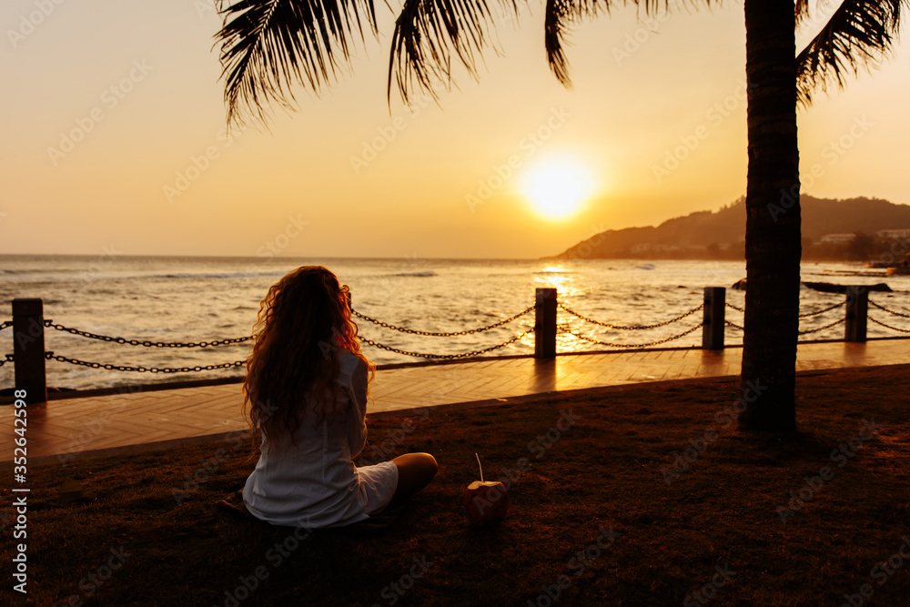 Back view of young lady with curly hair in white shirt sitting on ground with crossed legs and looking at beautiful sunset over sea. Happy woman enjoying perfect beach holiday vacation. Back view of young lady with curly hair in white shirt sitting on ground with crossed legs and looking at beautiful sunset over sea. Happy woman enjoying perfect beach holiday vacation.