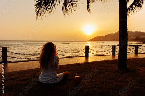 Back view of young lady with curly hair in white shirt sitting on ground with crossed legs and looking at beautiful sunset over sea. Happy woman enjoying perfect beach holiday vacation. © Tymoshchuk