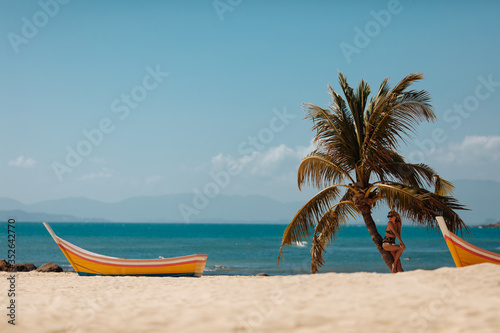 Charming girl with curly blond hair standing under palm tree leaves on sandy beach. Young woman in swimsuit enjoying exotic vacation near sea. Concept of travelling.
